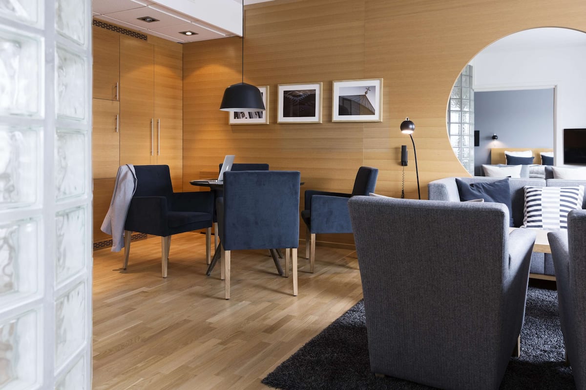 A modern hotel room with a wooden floor, a dining area with blue chairs, a sofa with striped pillows, and a large mirror.
