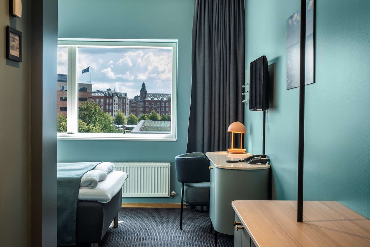 A hotel room with a bed, a desk, a chair, a lamp, a TV, and a window with a city view.