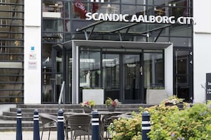 The exterior of Scandic Aalborg City hotel with a modern glass facade, outdoor seating area, and a sign indicating handicap accessibility.