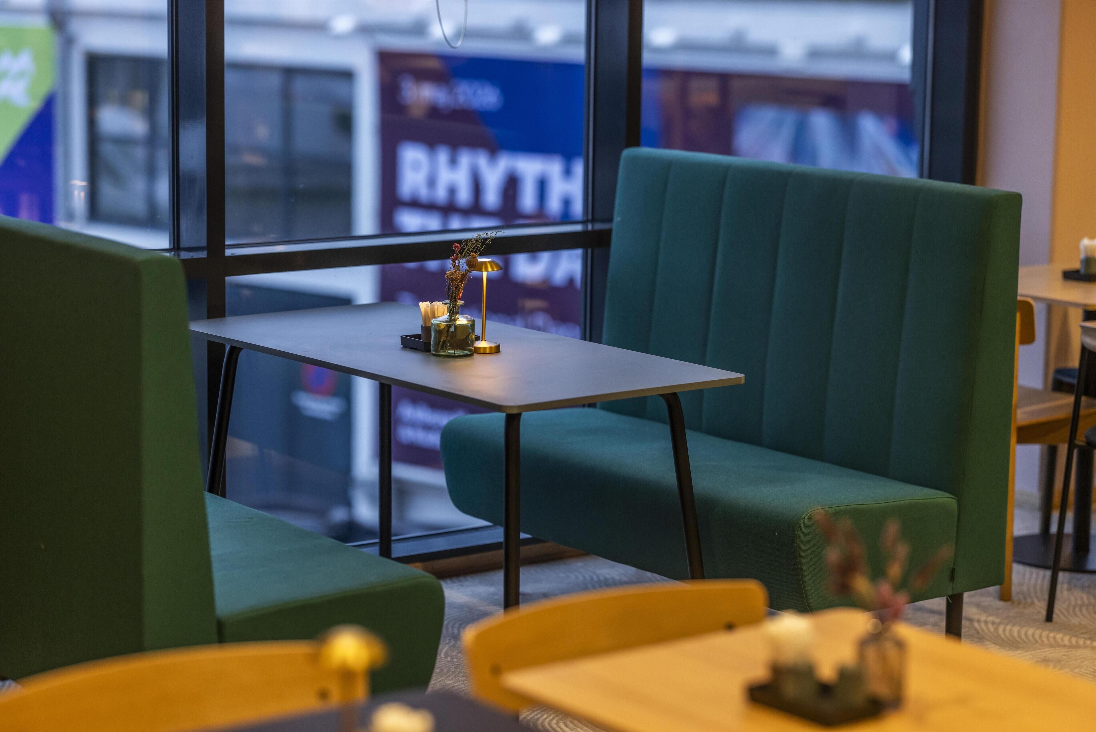 Uma área de estar moderna com cadeiras estofadas em verde e uma pequena mesa, com um vaso decorativo e uma luminária.