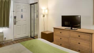 Quarto de hotel com uma cama, uma televisão sobre uma cômoda de madeira, um tapete verde e branco, uma porta com espelho e uma luminária fixada na parede.