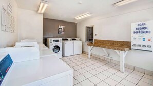 A laundry room with multiple washing machines, a bench, and a sign listing soap, bleach, and softeners. - Studio 6 Extended Stay - Ocean Springs, MS (Ocean Springs)