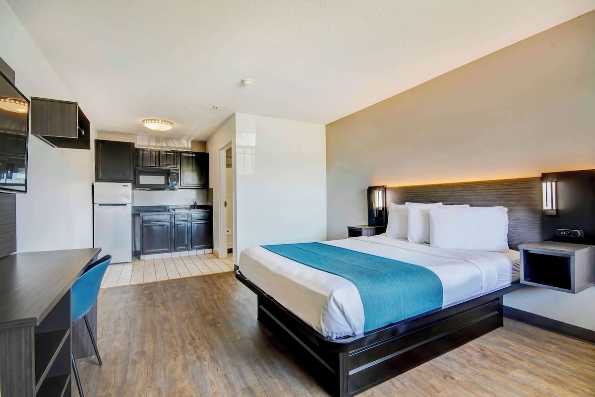 Una habitación de hotel moderna con cama, mesita de noche y un pequeño escritorio con silla. Cuenta con una kitchenette equipada con microondas y refrigerador.