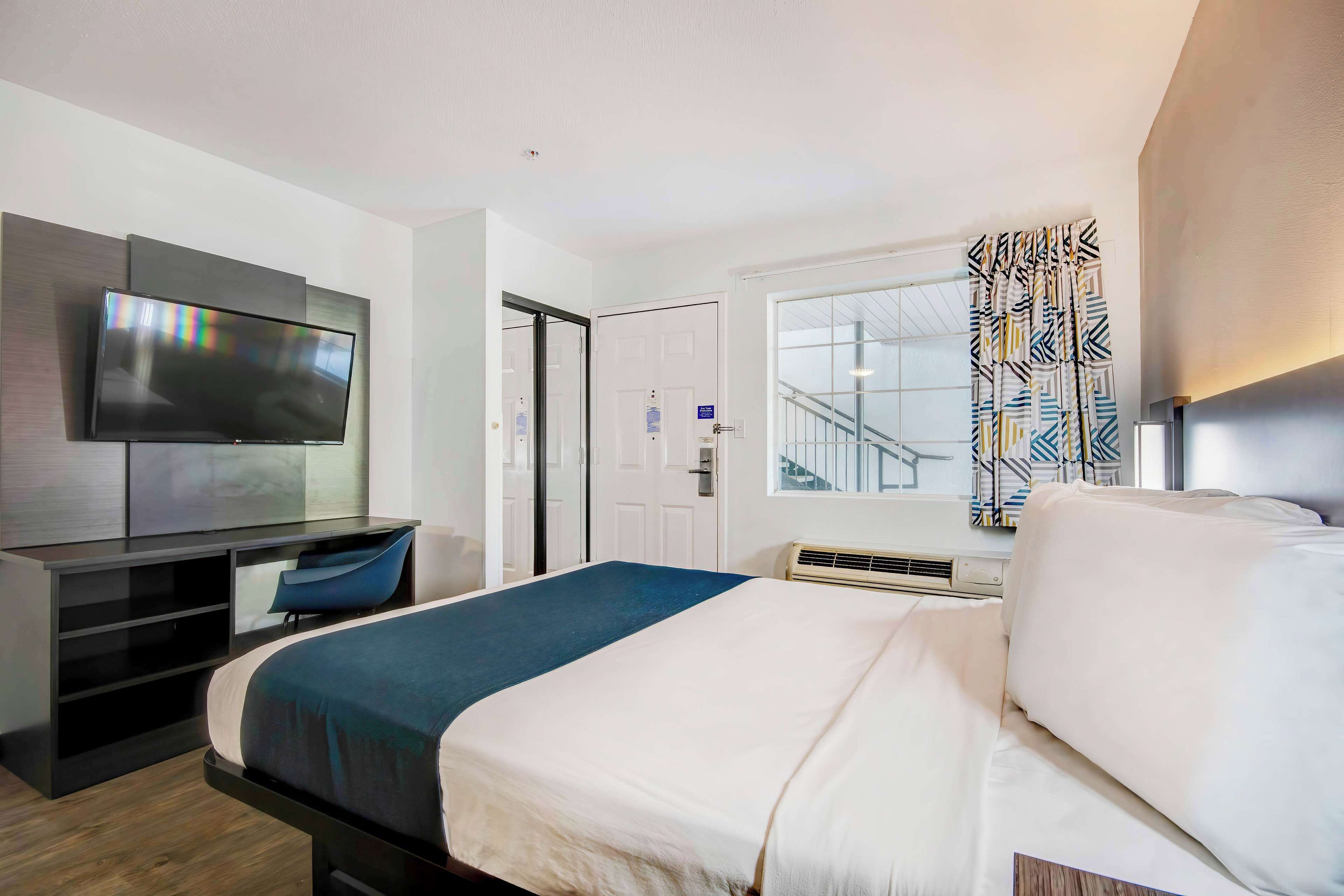 A modern hotel room with a bed, a desk with a chair, a flat-screen TV, a window with patterned curtains, and a staircase visible through the door.