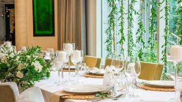 A dining table set with white tablecloths, candles, and glassware, surrounded by chairs with beige upholstery.