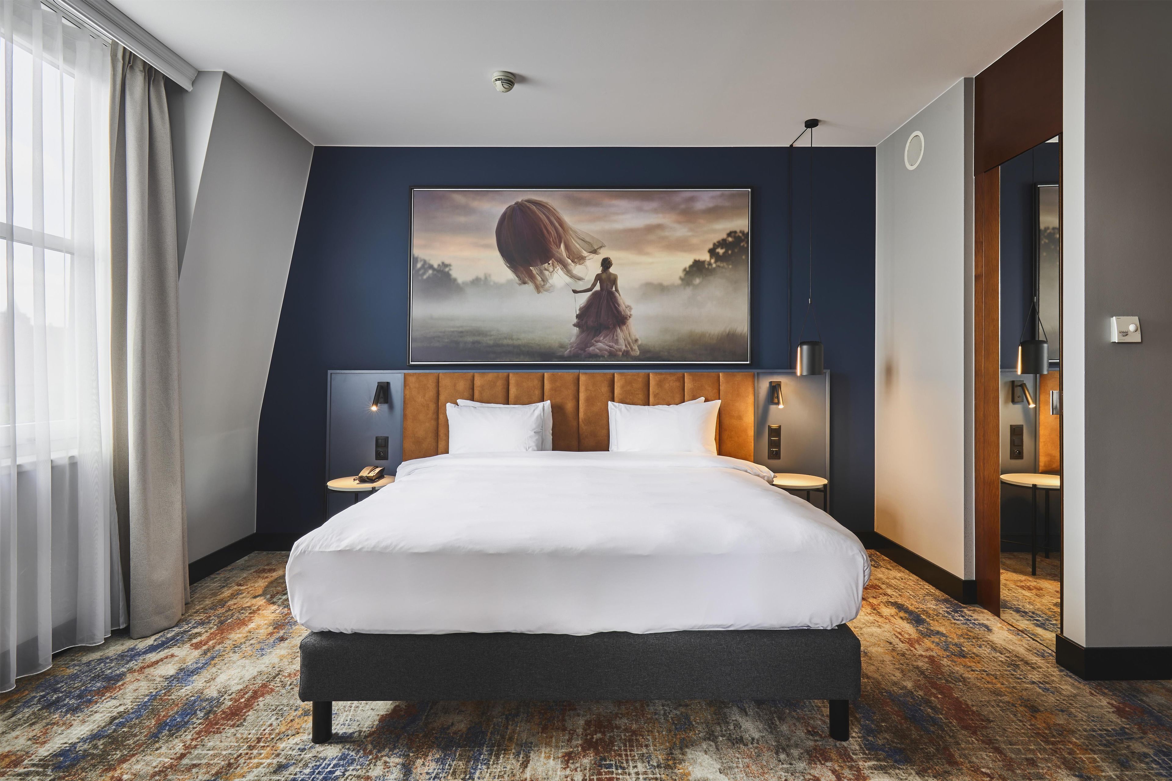 Habitación de hotel con una cama grande, cabecera, mesitas de noche, un cuadro y vistas al exterior.