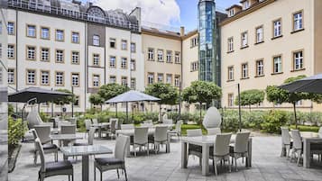 Eine Außenpatio-Terrasse mit Korbmöbeln und Sonnenschirmen, umgeben von Gebäuden mit zahlreichen Fenstern und Balkonen.