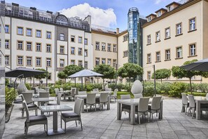 Un patio al aire libre con mobiliario de mimbre y sombrillas, rodeado de edificios con múltiples ventanas y balcones.