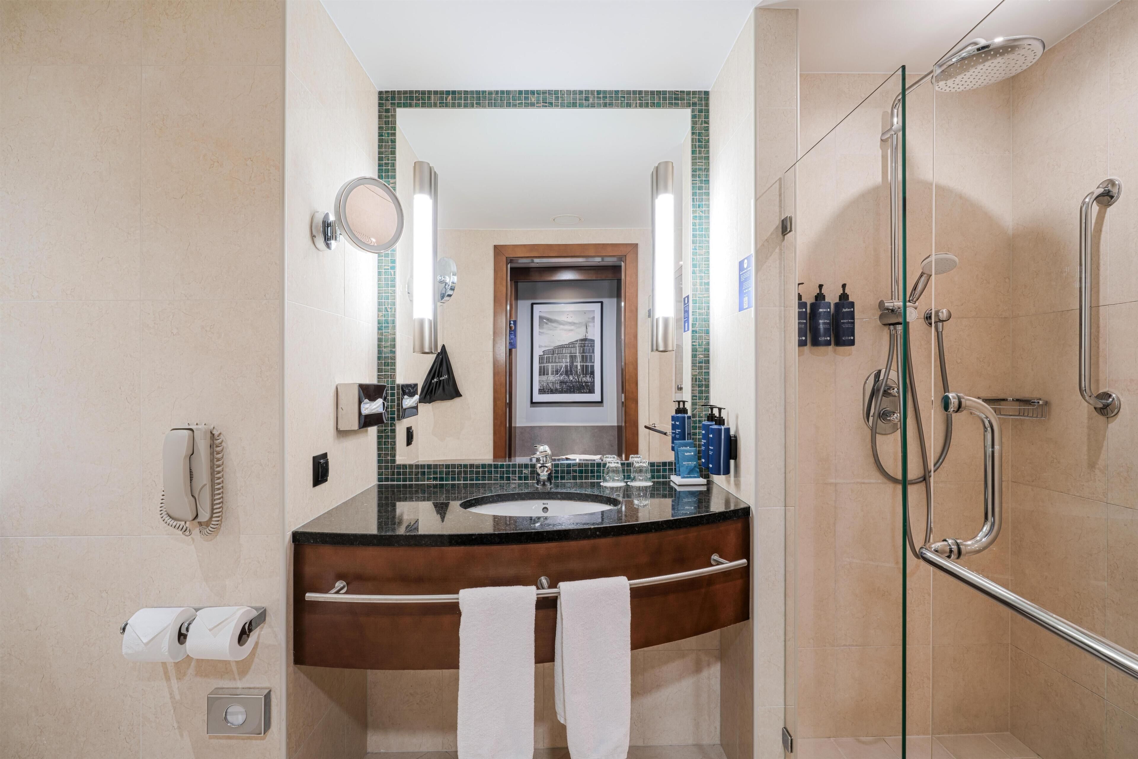 A bathroom with a sink, mirror, towel rack, shower, and toilet paper dispenser.