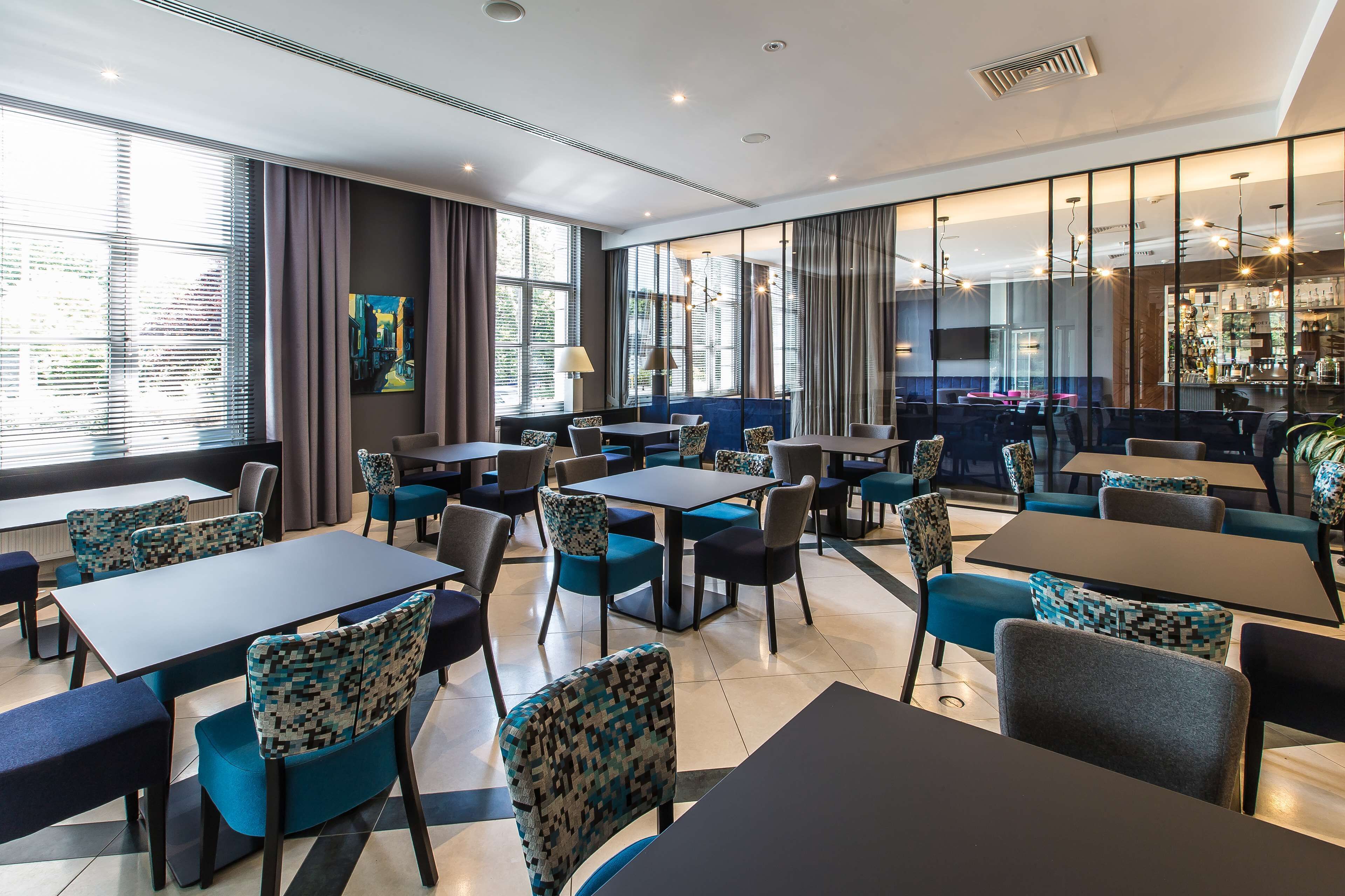 A modern hotel dining area with multiple tables and chairs, large windows with curtains, and a bar area in the background.