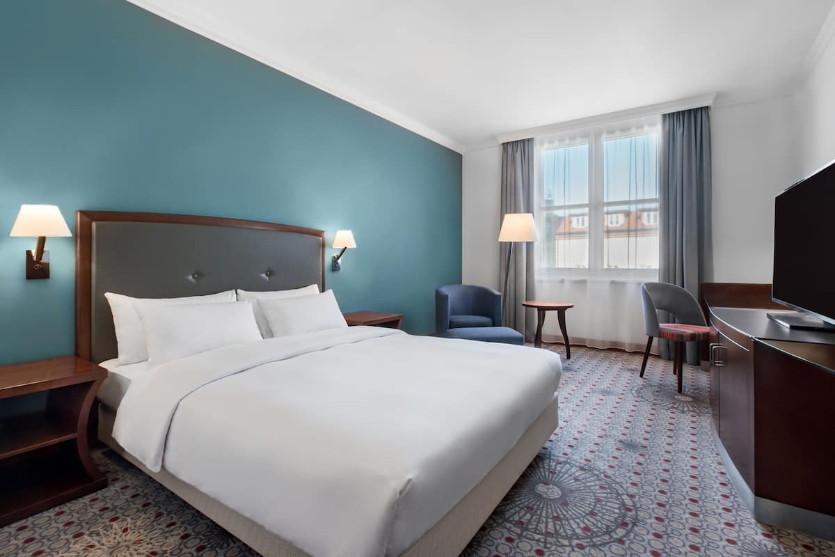 Habitación de hotel con una cama grande, dos mesitas de noche con lámparas, un escritorio con una silla, un televisor y una ventana con cortinas.