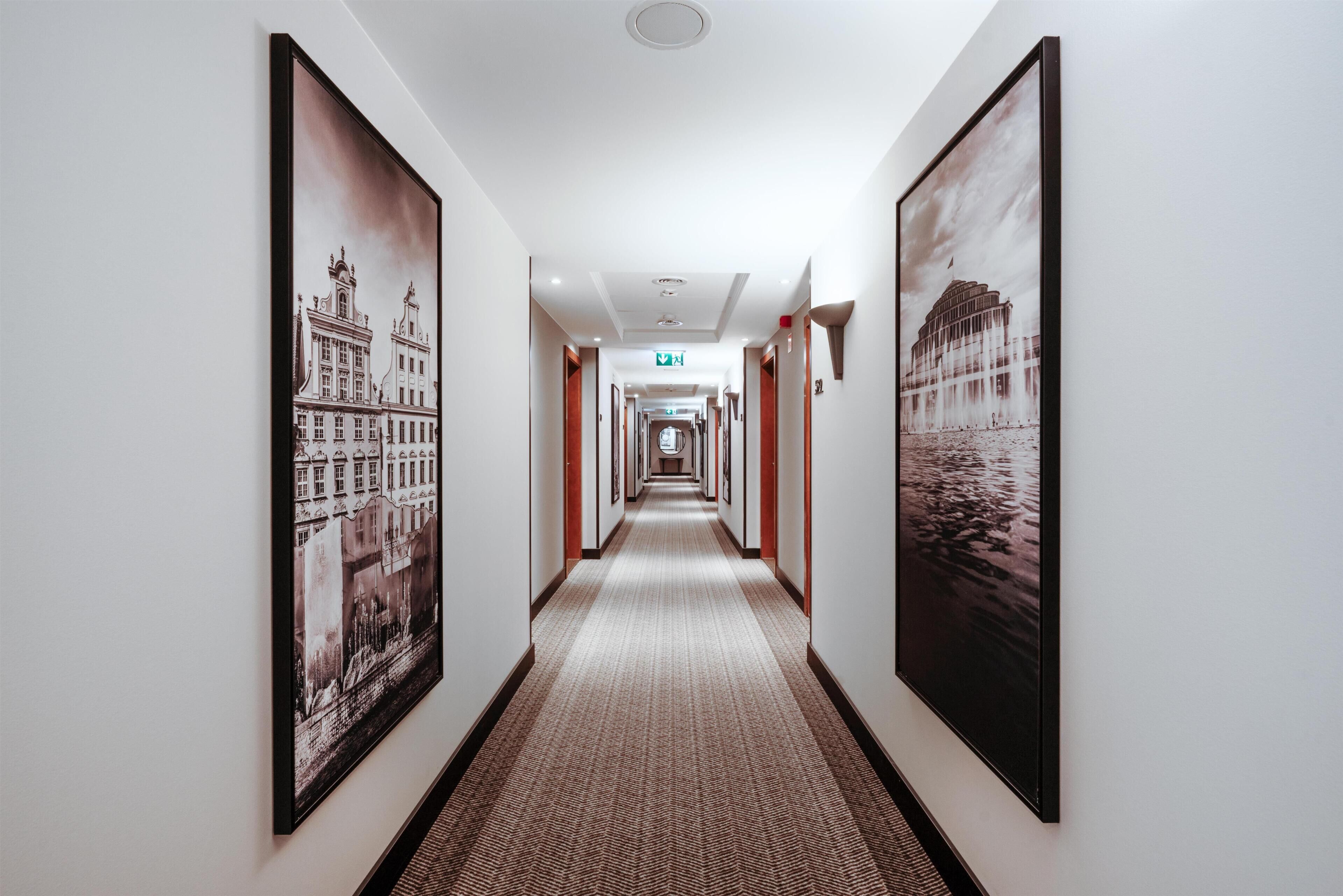 Corredor de hotel com piso de carpete, quadros nas paredes e sinalização de saída.