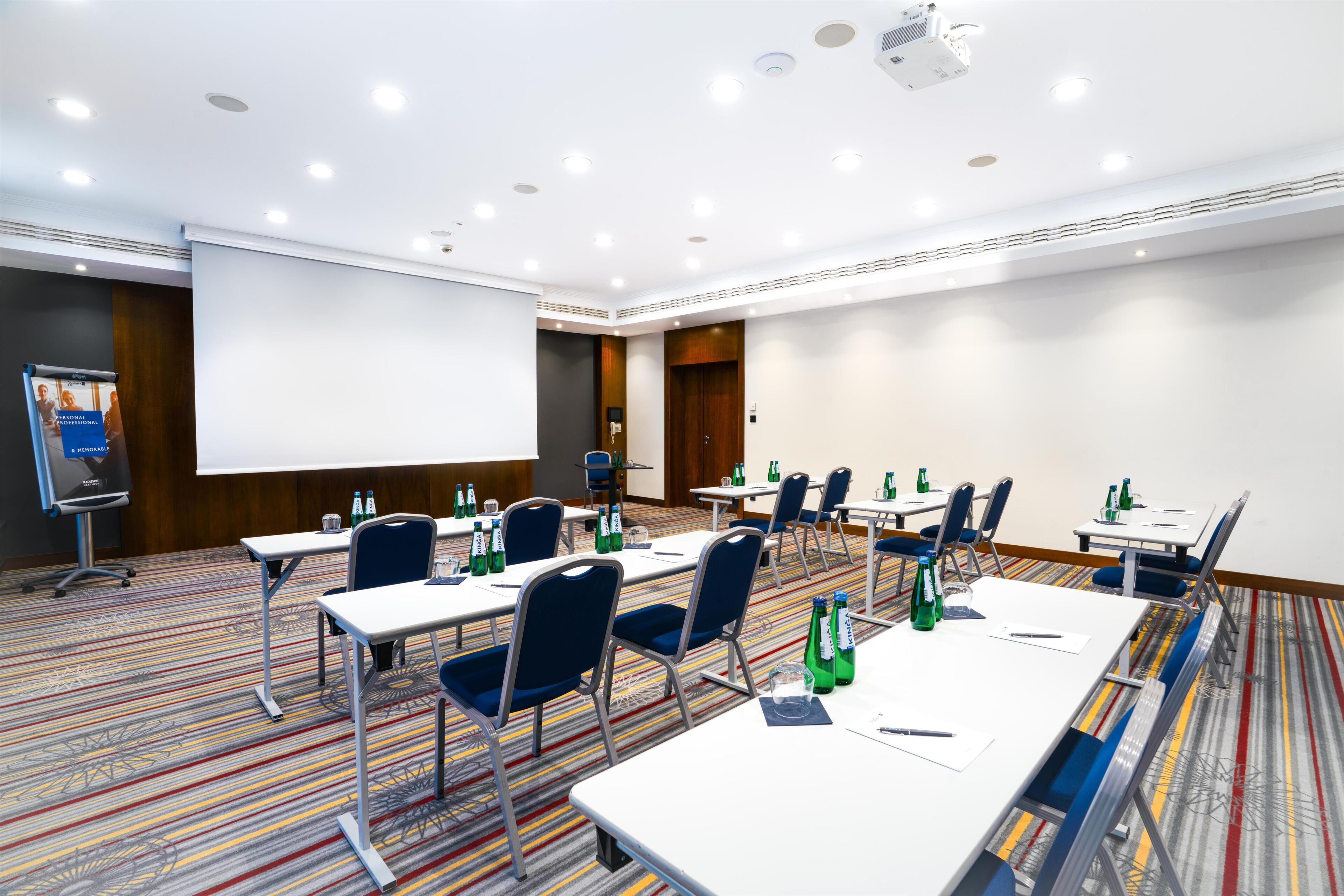 Una sala conferenze con tavoli, sedie, uno schermo per proiettore e pavimento con moquette.