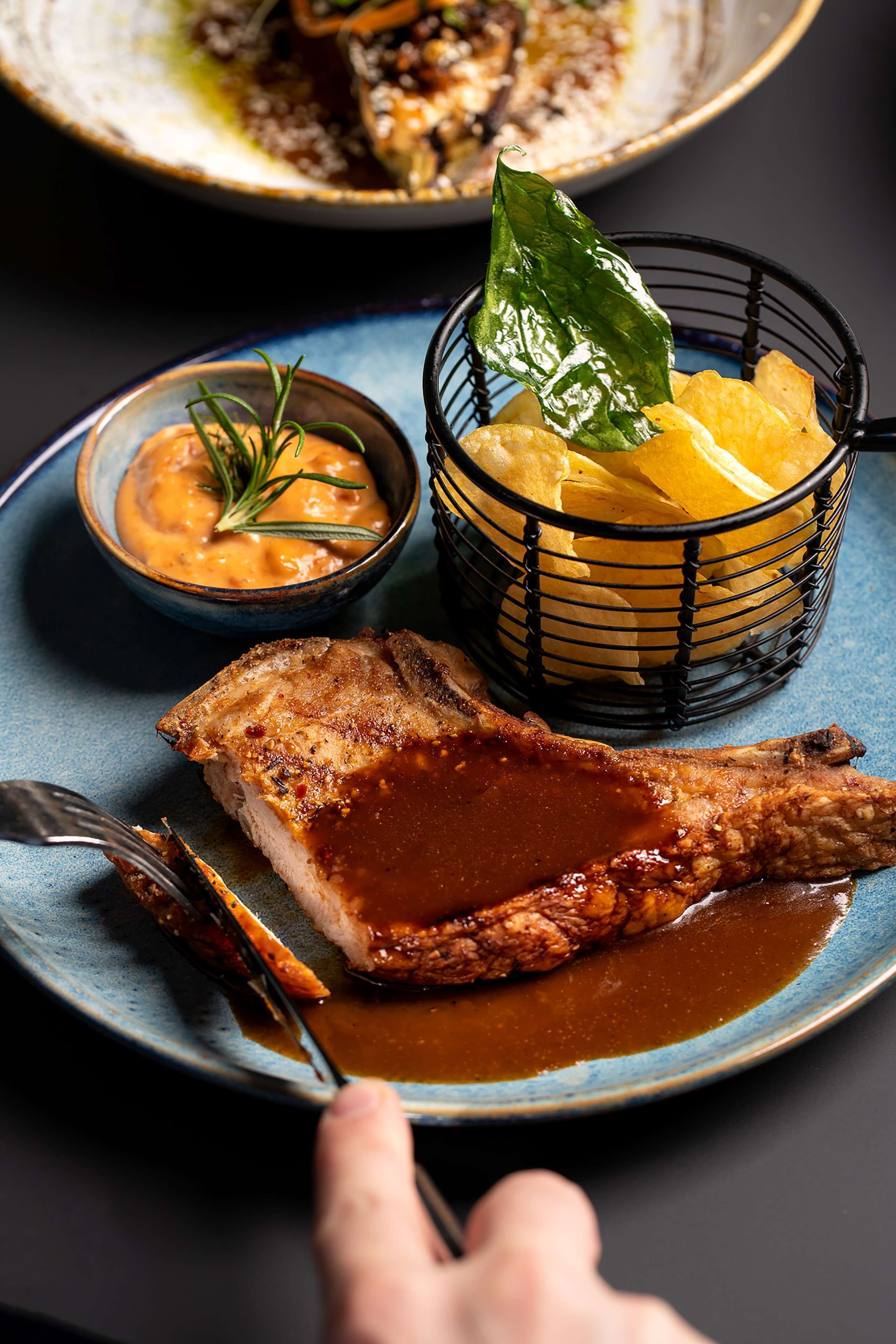 A plate with a piece of meat, a bowl of sauce, and a basket of chips.