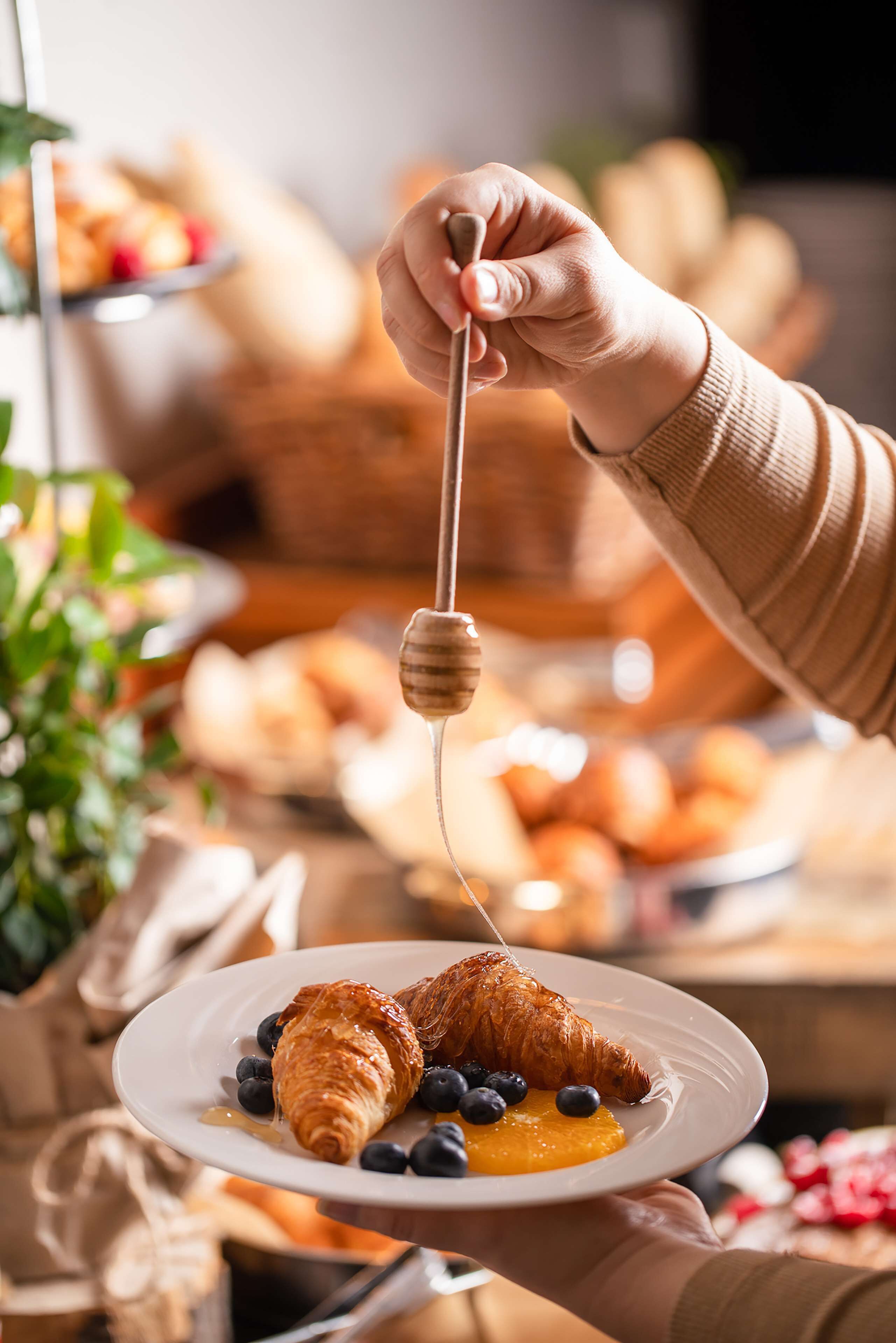A hand holding a honey dipper over a plate with a croissant, blueberries, and orange slices.