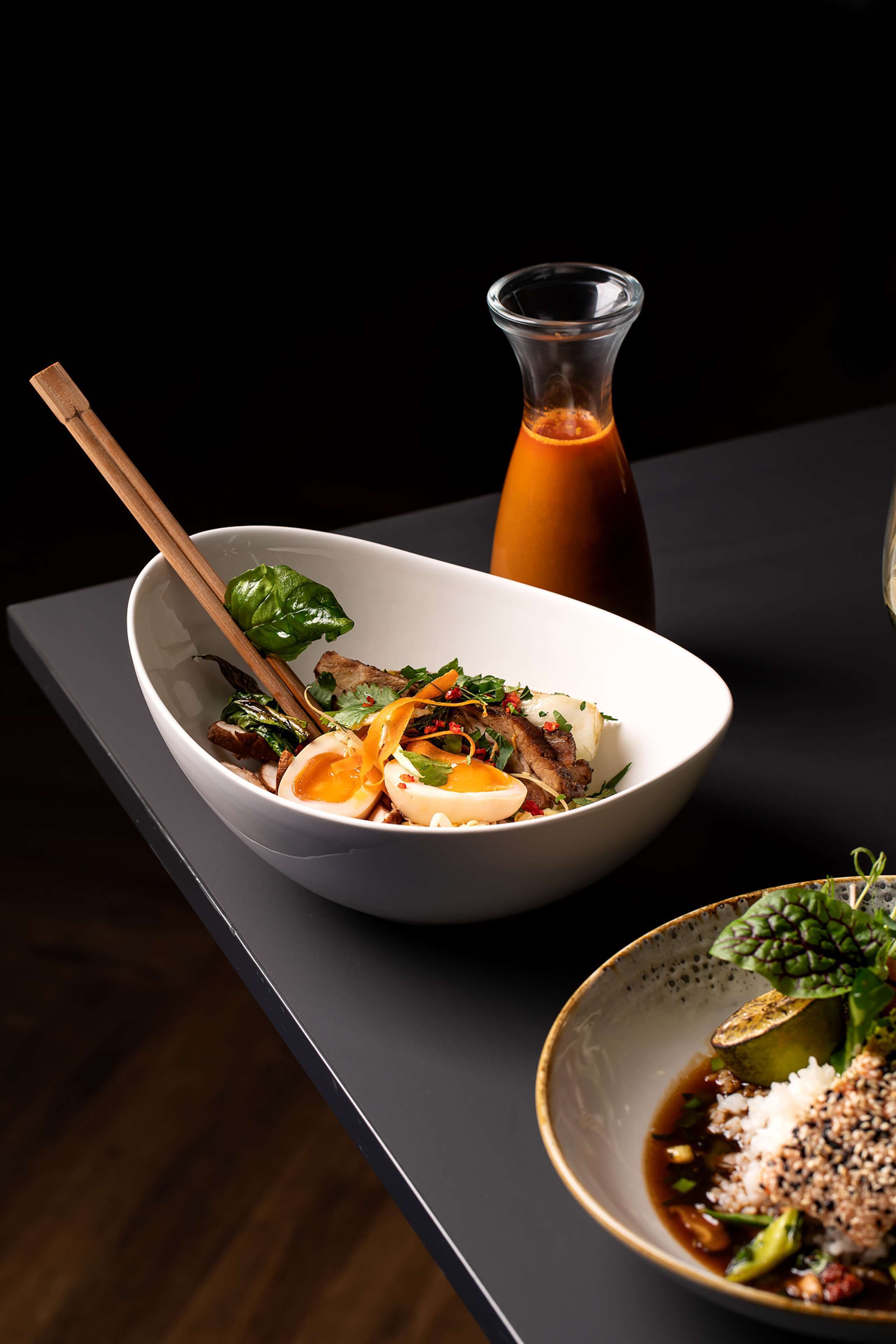 A bowl of food with chopsticks, a bottle of sauce, and a glass of water on a table.