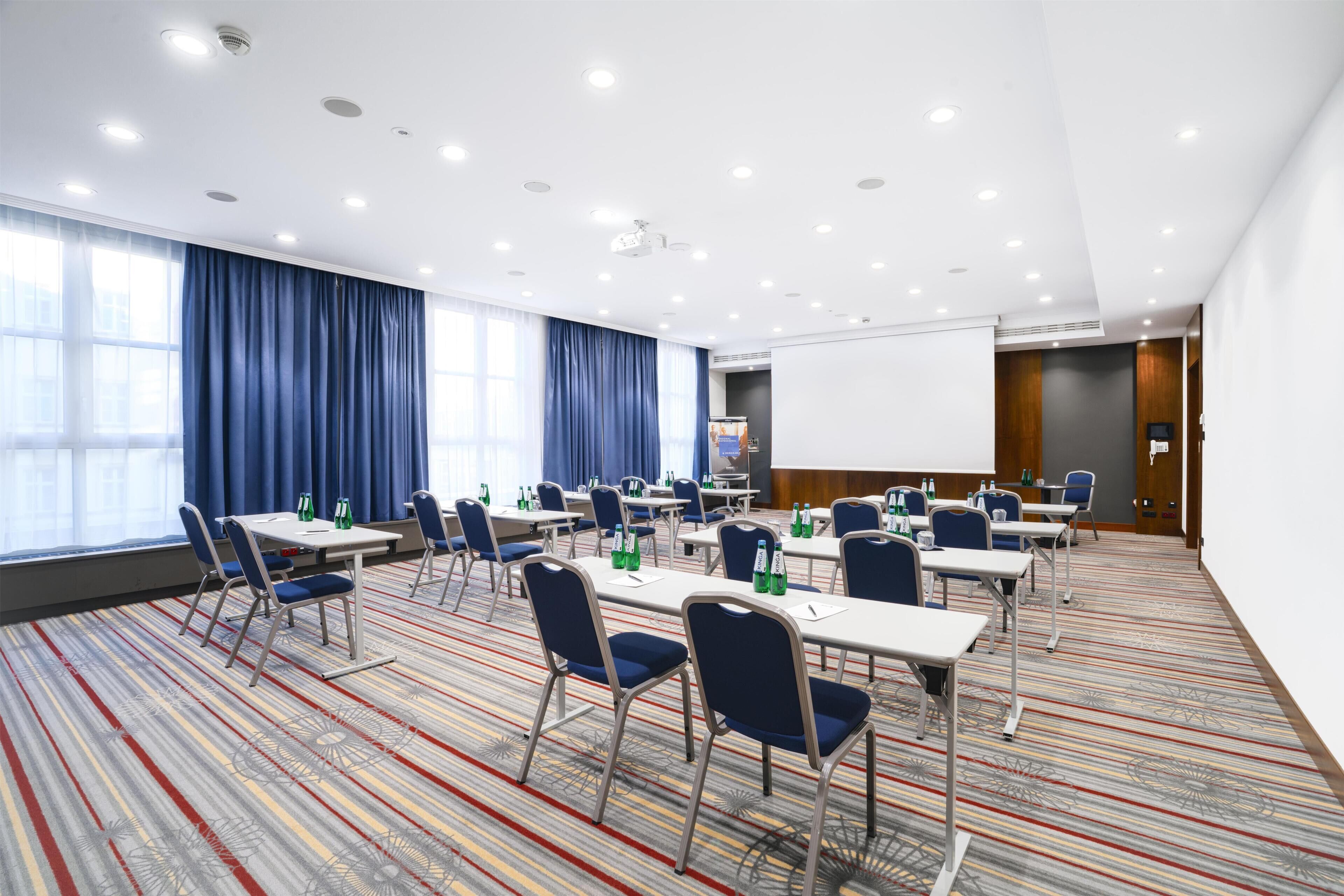 A conference room with tables, chairs, a projector screen, and a large window with curtains.