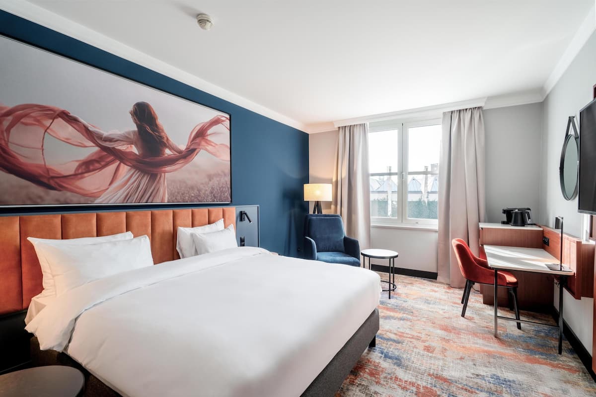 Habitación de hotel con una cama grande, un escritorio, una silla, una lámpara y una ventana con cortinas.