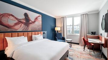Quarto de hotel com uma cama grande, uma escrivaninha, uma cadeira, um abajur e uma janela com cortinas.