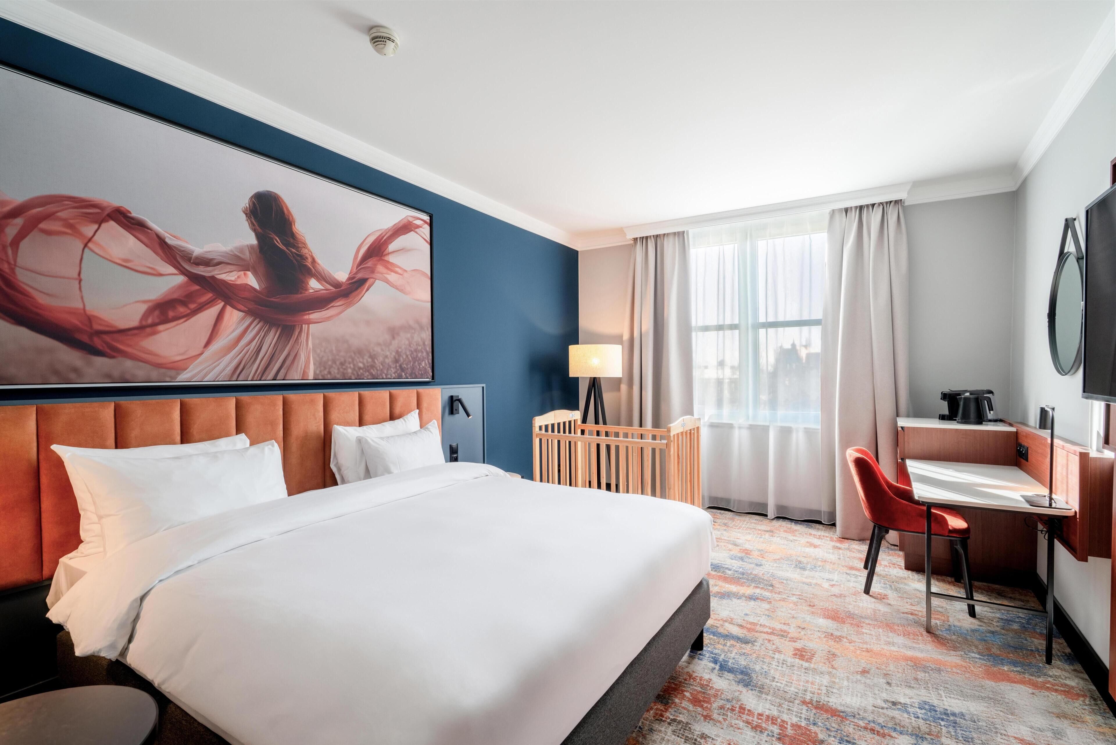 Una habitación de hotel con una cama grande, un escritorio, una silla, una lámpara y vistas a la ciudad desde la ventana.