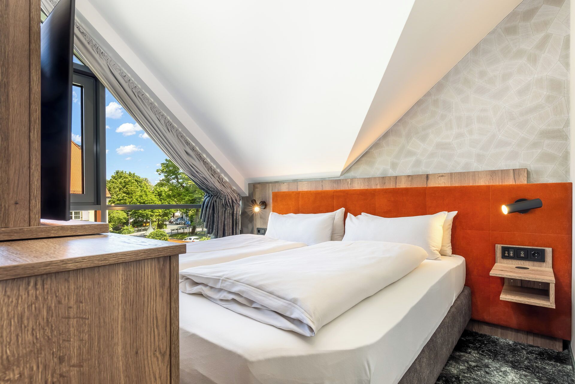 A hotel room with a bed, a TV, a wooden desk, and a view of the outdoors.