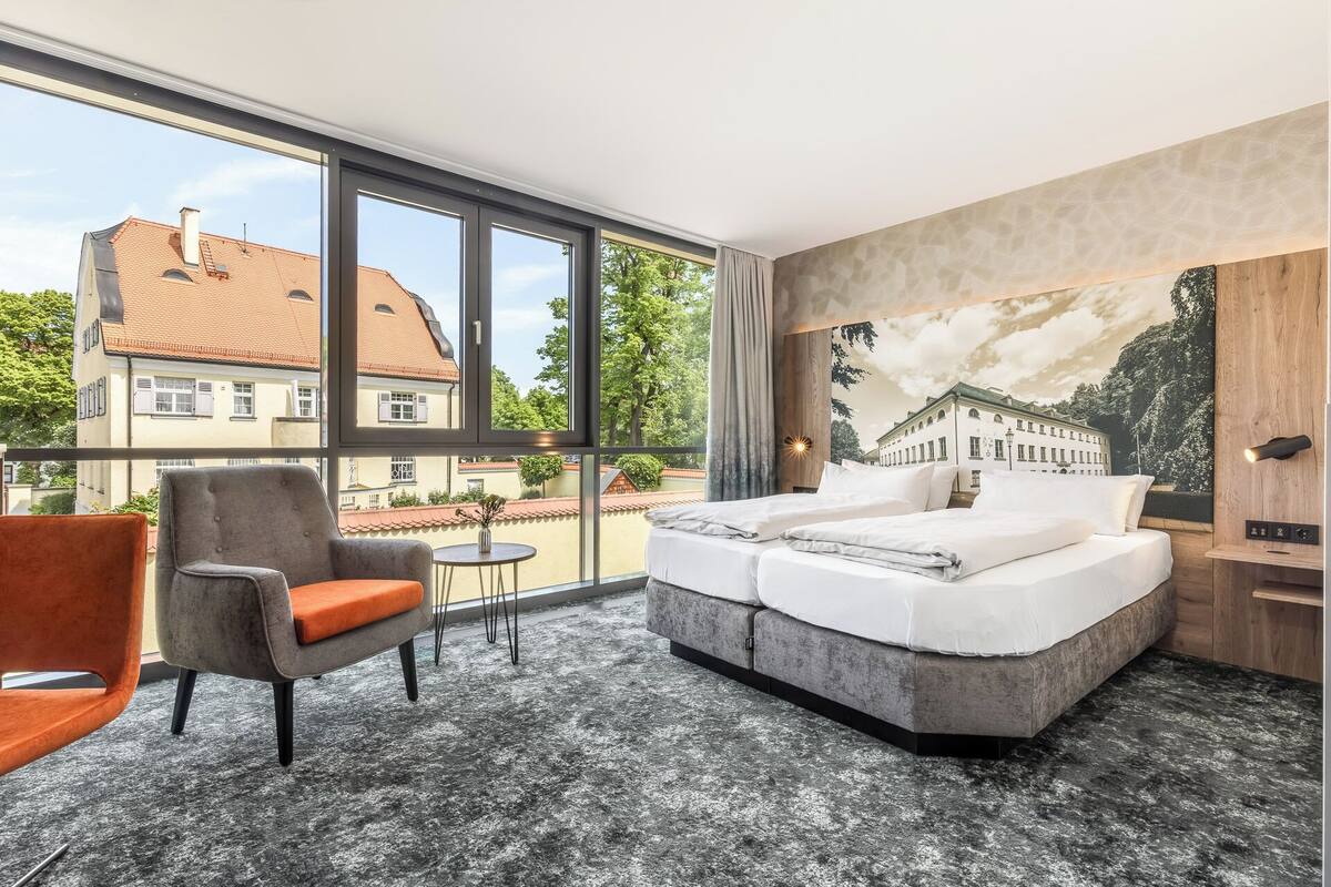 A hotel room with a large window, a bed, a chair, a small table, and a mural on the wall.