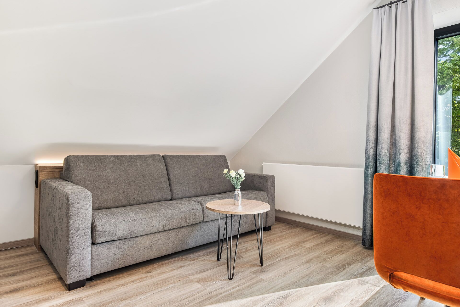 A modern loft bedroom with a grey sofa, a small round table with a vase of flowers, and a single orange chair.