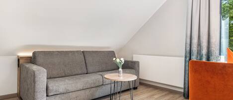 A modern loft bedroom with a grey sofa, a small round table with a vase of flowers, and a single orange chair.