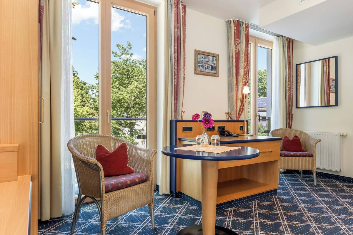 A room with a round table, wicker chairs, a mirror, and a view of trees through large windows.