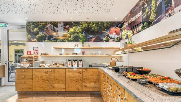 A buffet setup with a variety of food items, wooden cabinetry, and a large poster on the wall. - Hotel zur Post (Ismaning)