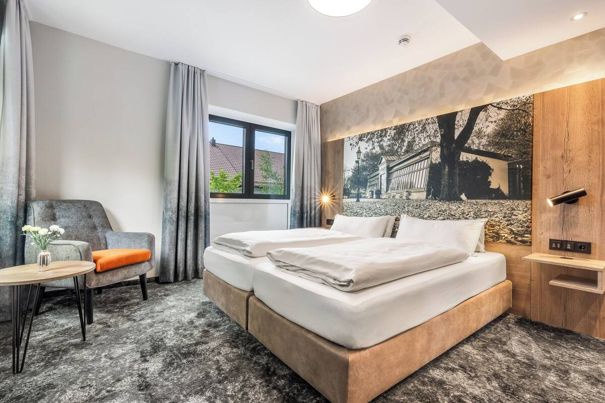 A hotel room with a bed, a chair, a small table, and a window with a view of buildings.