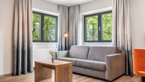 A modern living room with a grey sofa, a wooden coffee table, and a view of greenery through large windows. - Hotel zur Post (Ismaning)