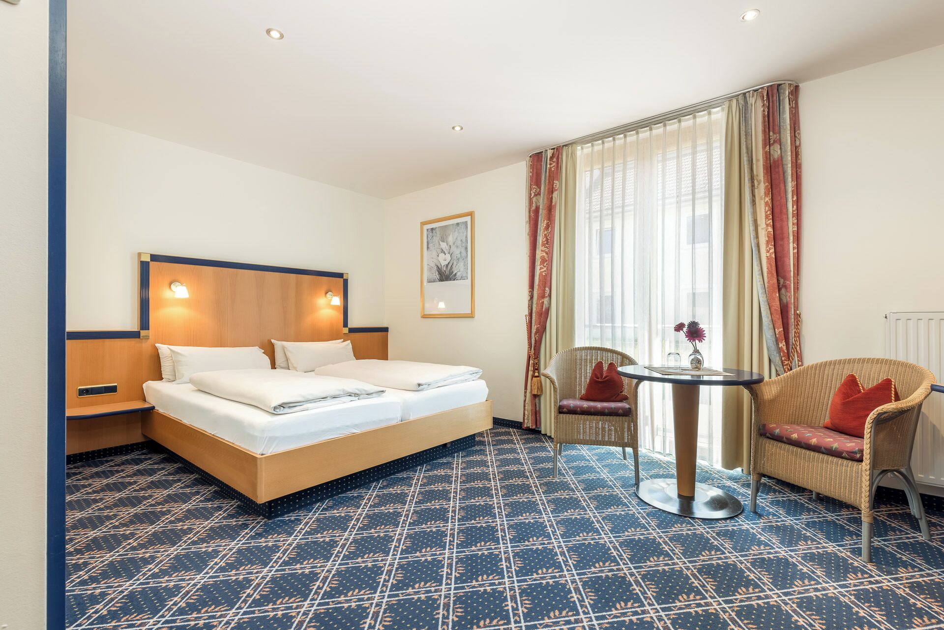 Habitación de hotel con una cama grande, dos mesitas de noche, una pequeña mesa redonda, una silla y un ventanal con cortinas.