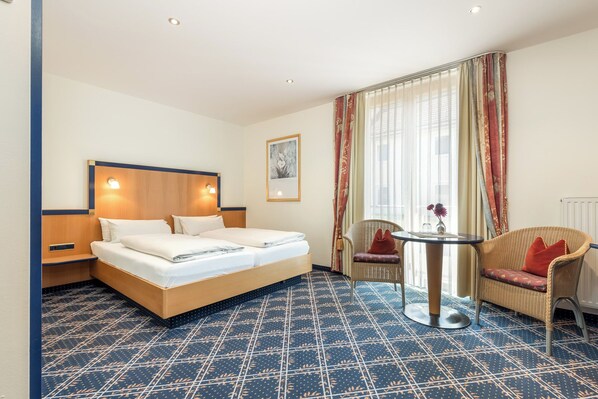 Habitación de hotel con una cama grande, dos mesitas de noche, una pequeña mesa redonda, una silla y un ventanal con cortinas.