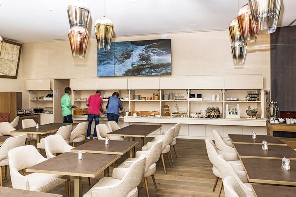 Un restaurante con varias mesas y sillas, una zona de cocina con personal, y un cuadro grande colgado en la pared que muestra un río.