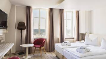 Ein Hotelzimmer mit zwei Betten, einem Schreibtisch, einem Sessel, einem Fernseher und Meerblick.
