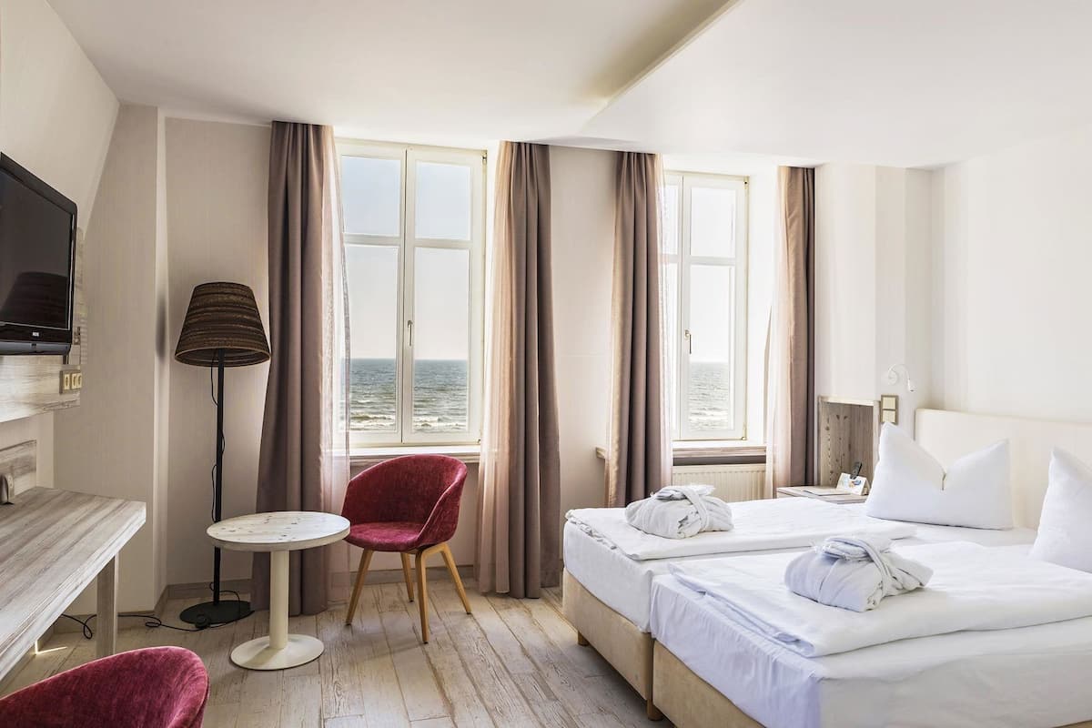 A hotel room with a bed, a desk with a TV, a chair, a lamp, and a view of the sea.