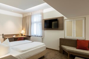 A hotel room with a bed, a desk, a TV, and a window with blinds.