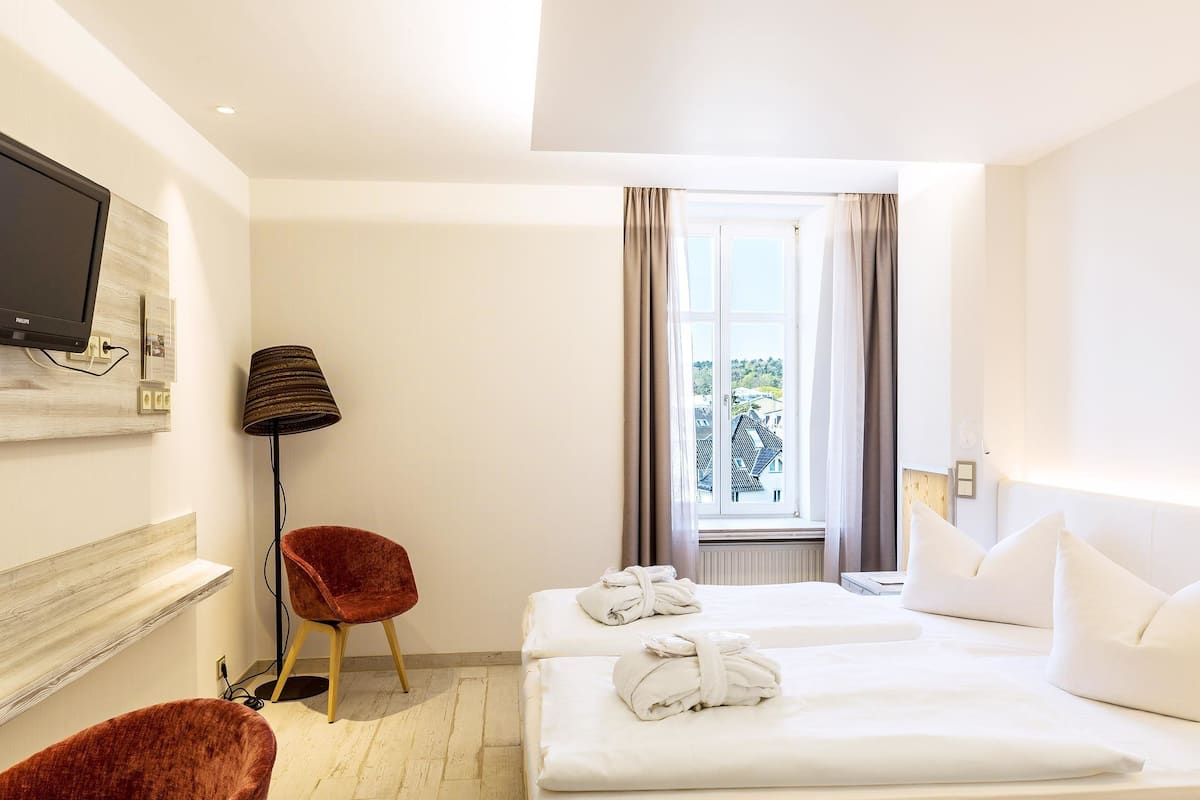 A hotel room with two beds, a TV mounted on the wall, a red chair, a floor lamp, and a window with curtains.