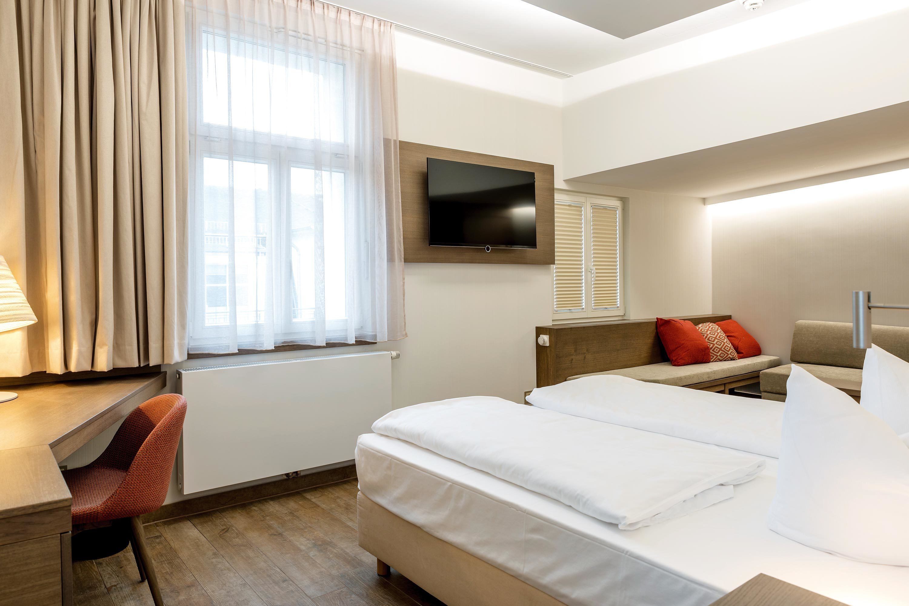 Une chambre d’hôtel avec un lit, un bureau, une chaise, une télévision et une fenêtre avec des rideaux.