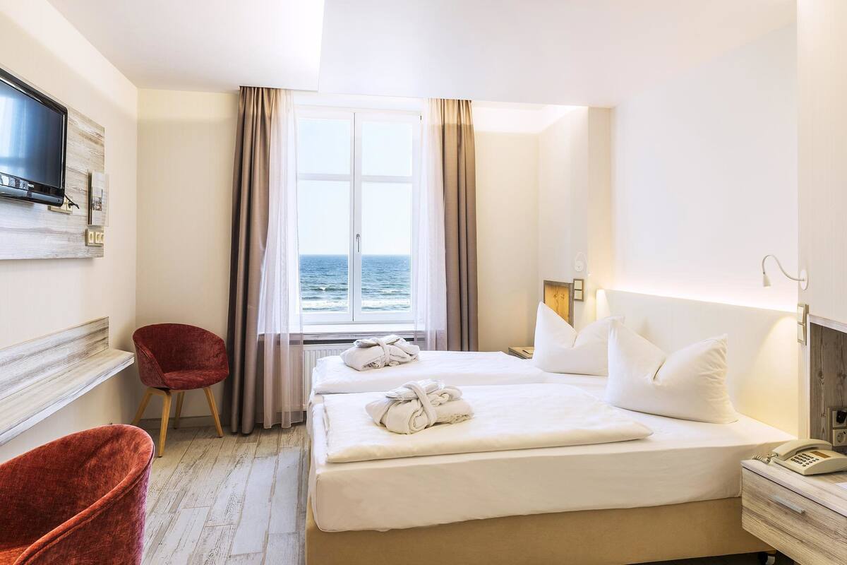 A hotel room with two beds, a TV mounted on the wall, a chair, and a view of the sea through the window.