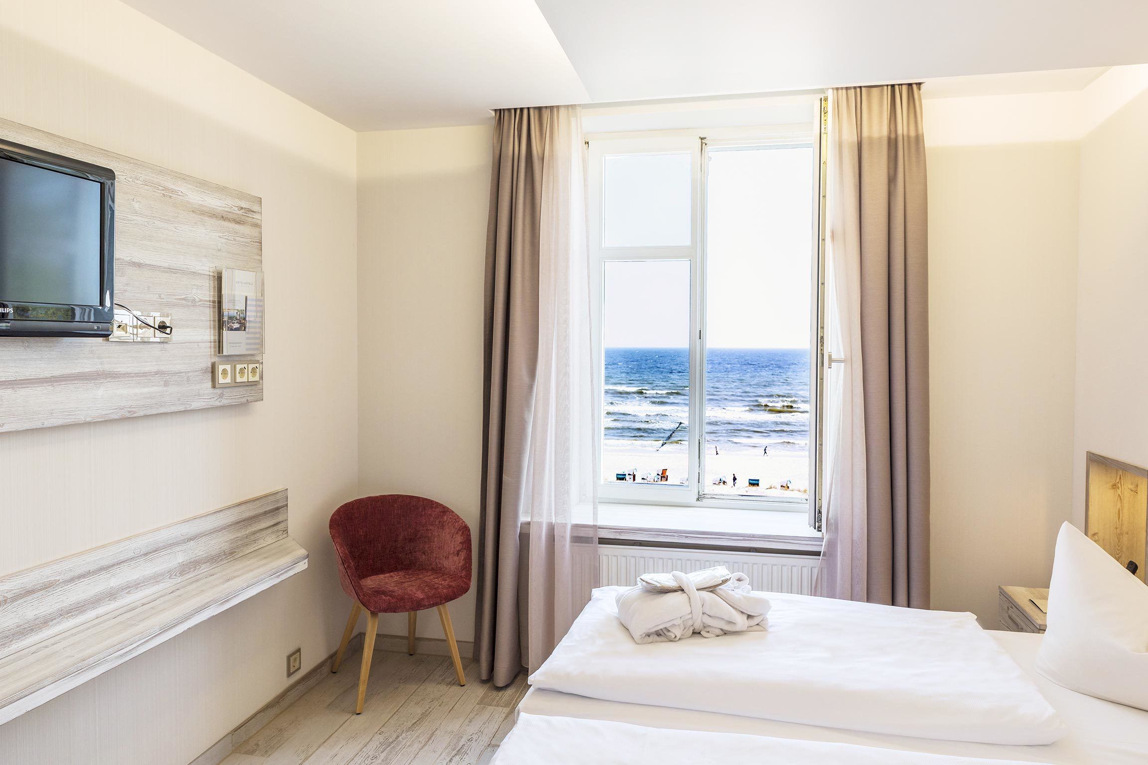 A hotel room with a bed, a red chair, a TV mounted on the wall, and a window with a view of the sea.
