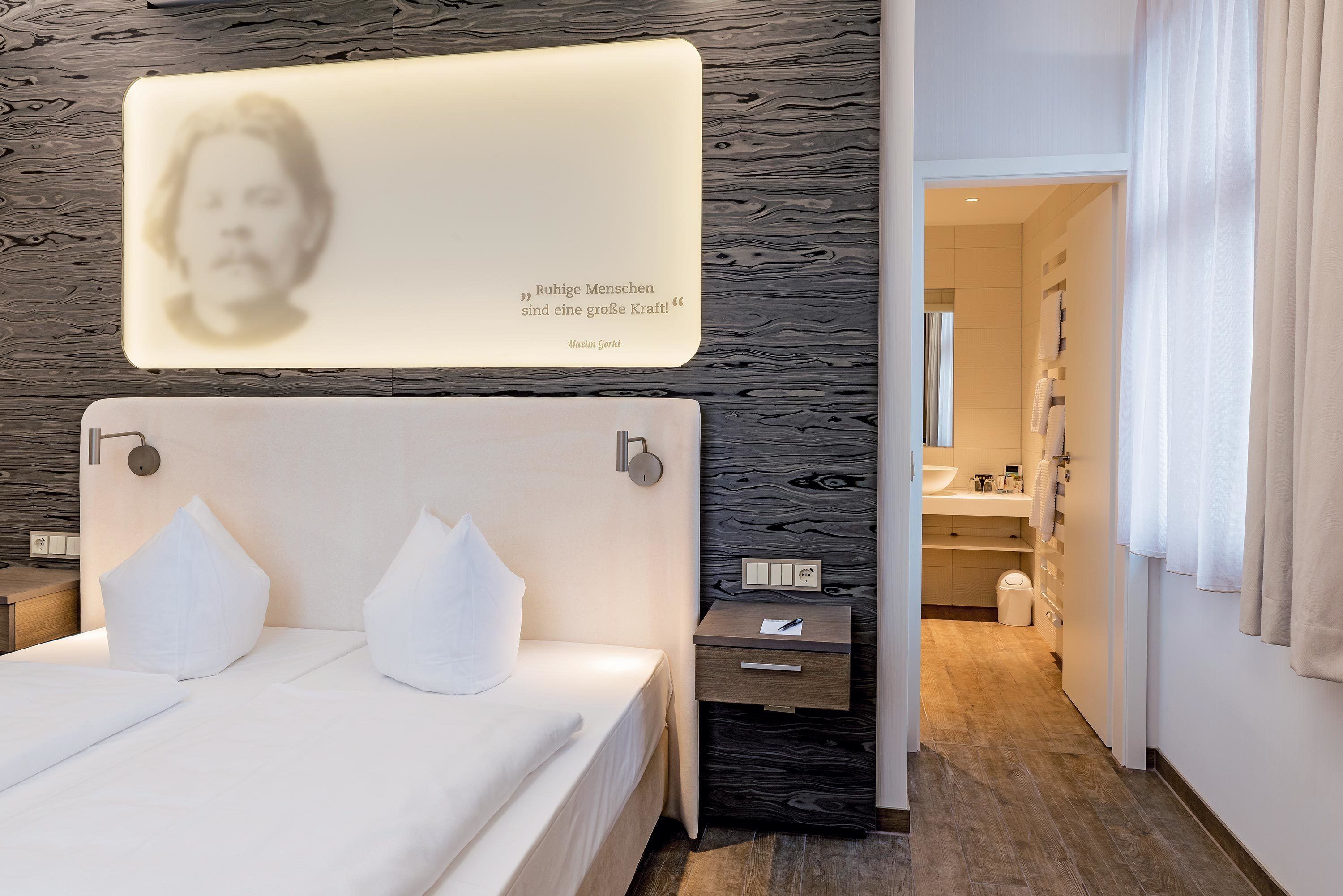 Una habitación de hotel moderna con una cama bien hecha, una mesita de noche con una lámpara y una lámpara de pared que muestra una cita.