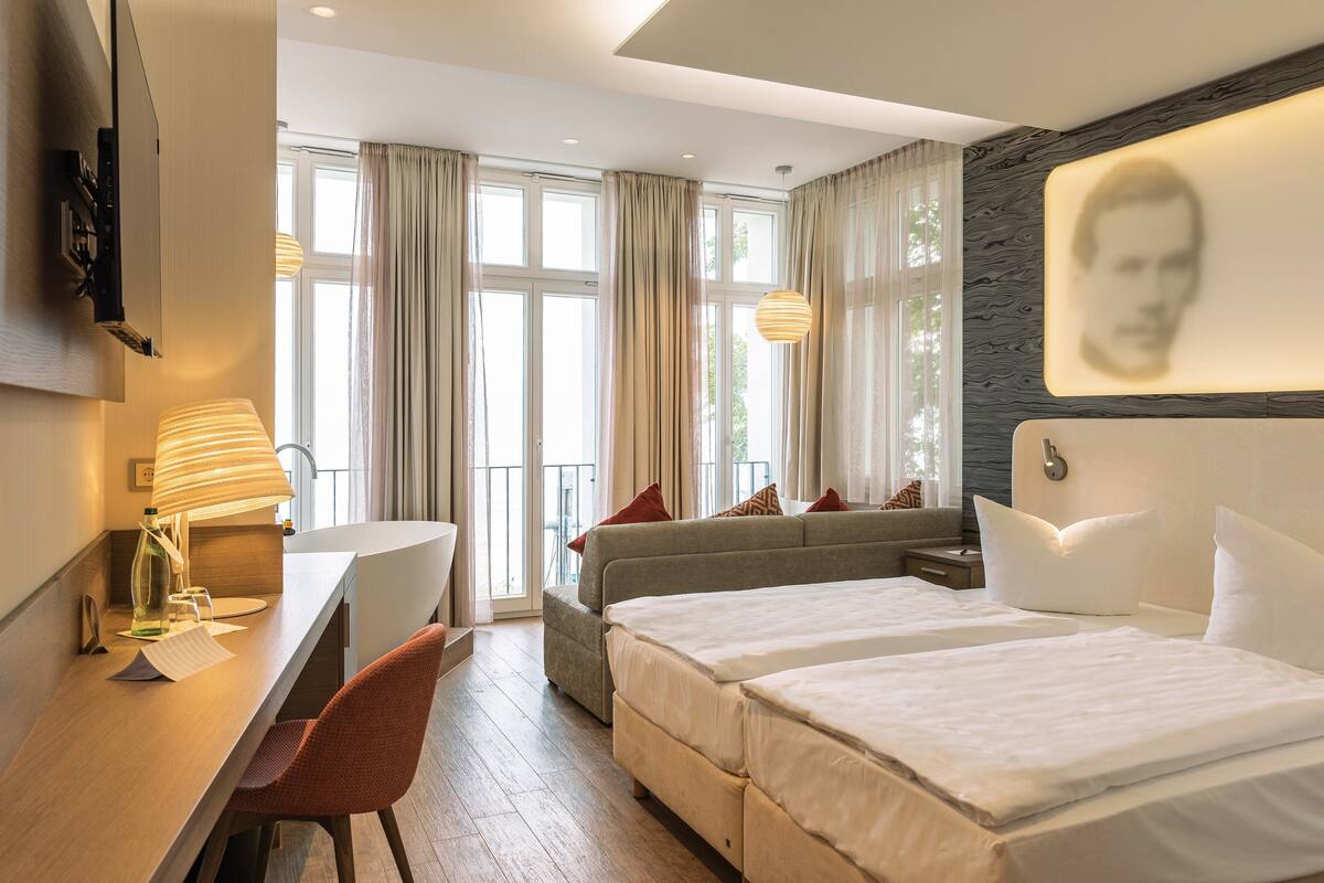 A hotel room with a desk, two beds, a sofa, and a large window with curtains.