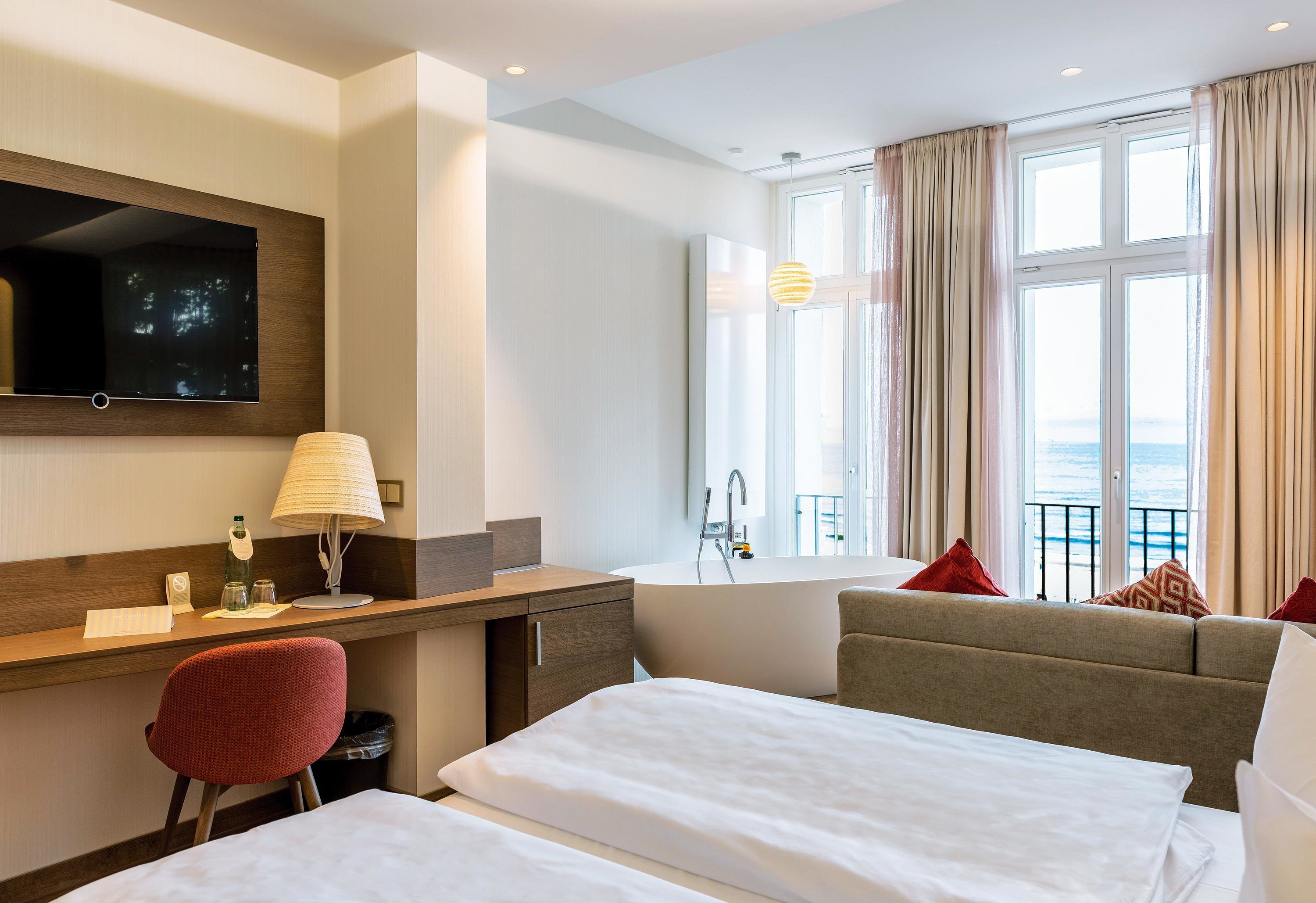A hotel room with a bed, a desk, a chair, a TV, a window with curtains, and a view of the sea.
