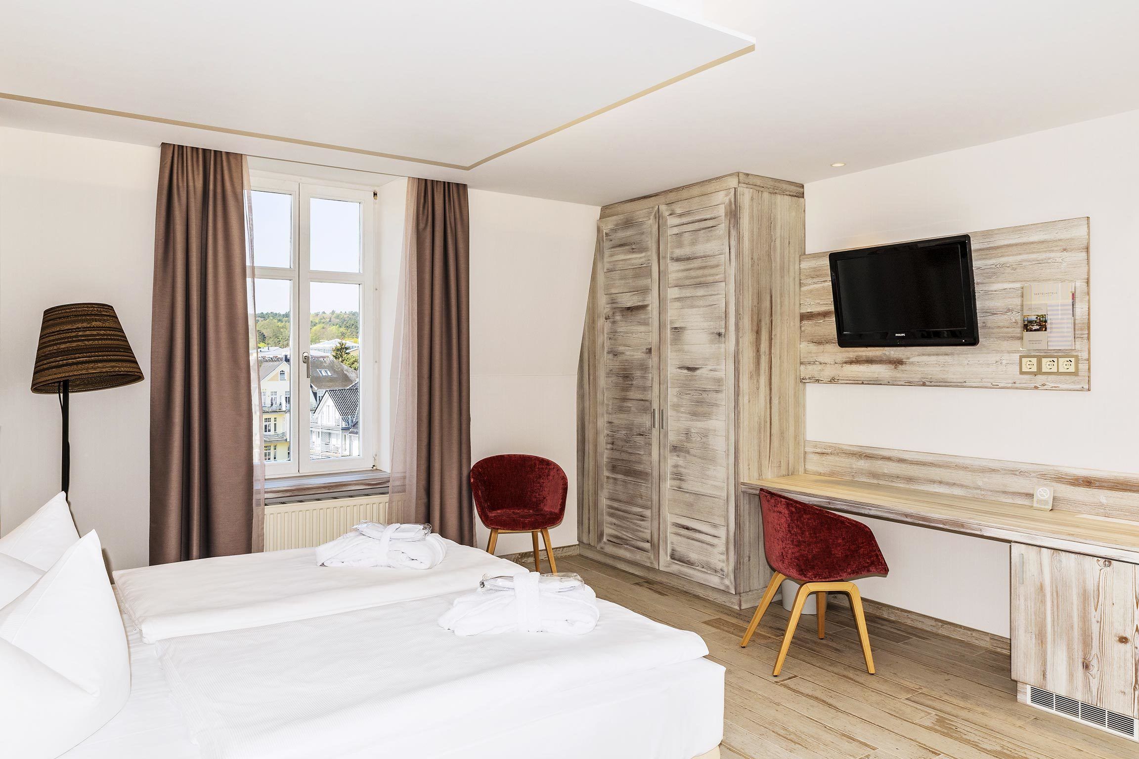 A hotel room with a bed, a desk, a TV, and a view of buildings outside.