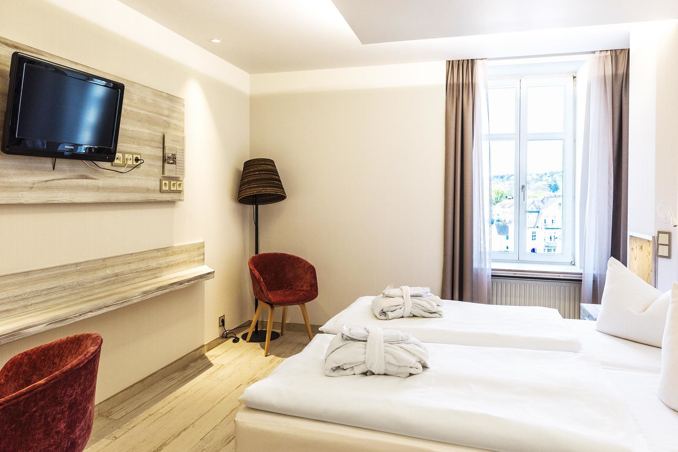 A hotel room with two beds, a TV mounted on the wall, a red chair, and a window with curtains.