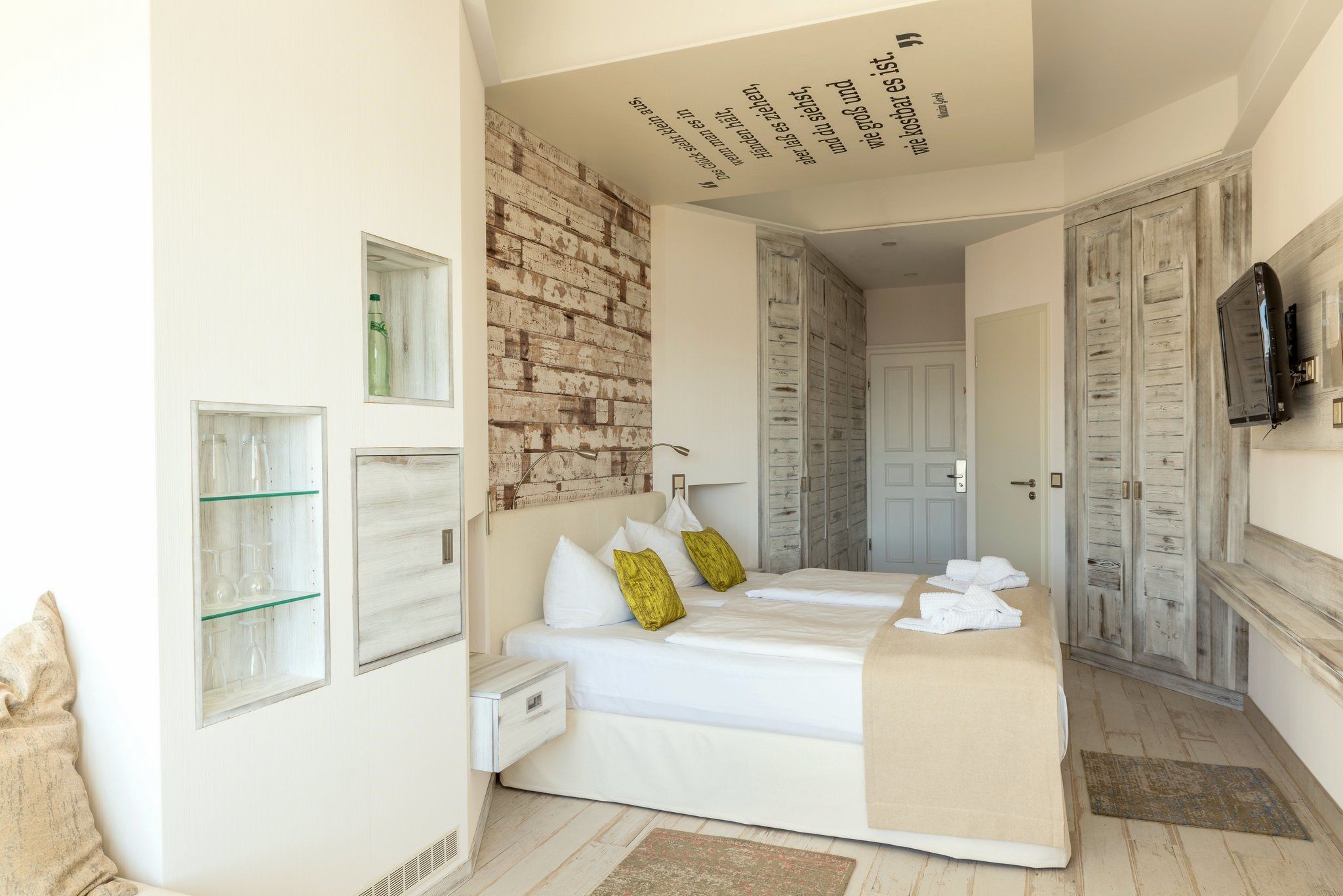 A modern bedroom with a brick accent wall, a bed with white bedding and green pillows, a bedside table, and a mirror.