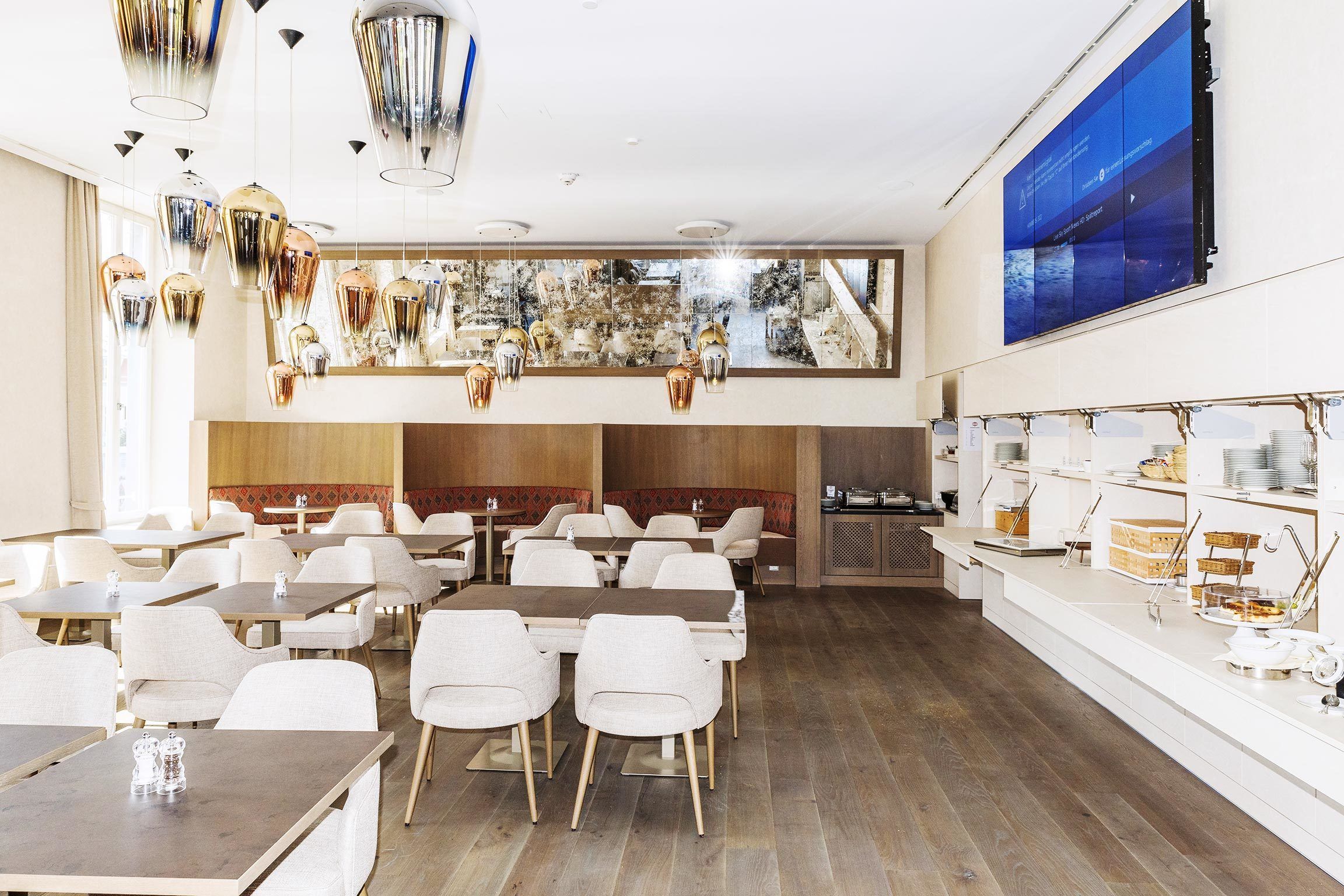 A modern dining area with multiple tables and chairs, a large digital display on the wall, and a view of the outside through large windows.