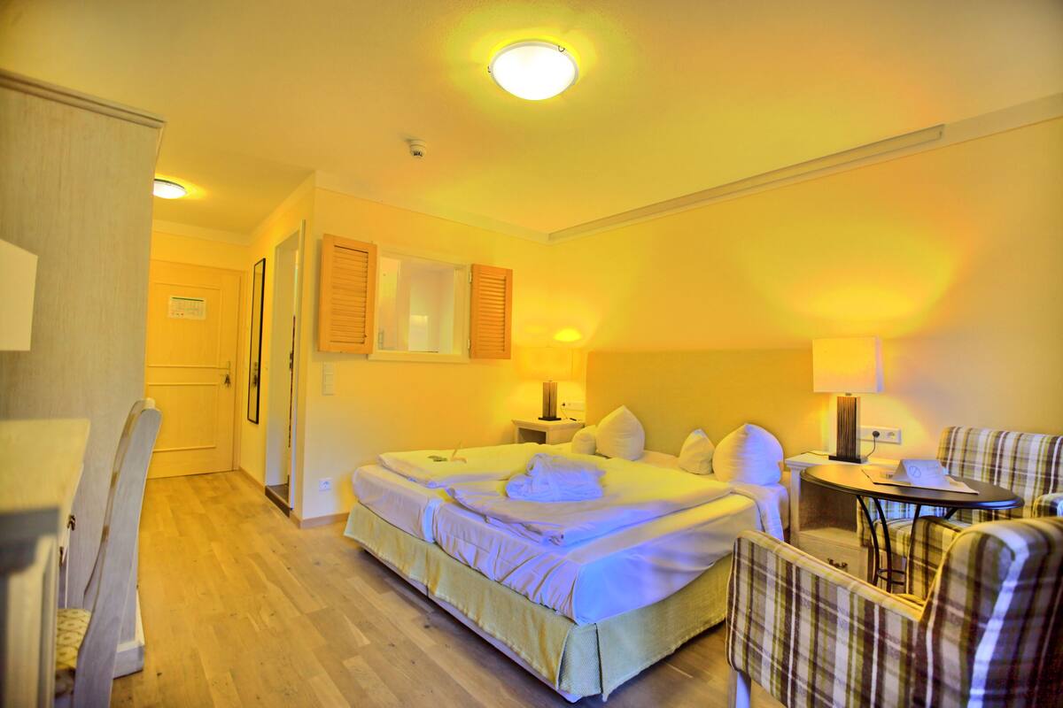 A hotel room with a large bed, a small table, two chairs, and a door leading to another room.